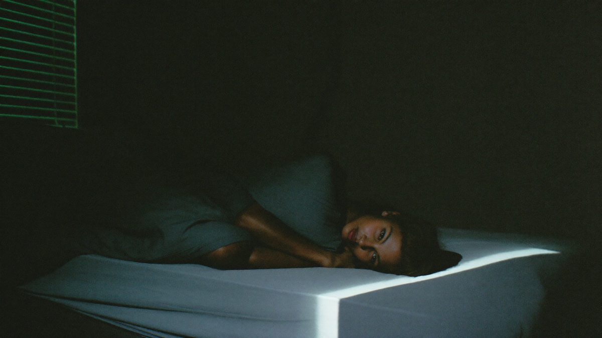 Person lying awake in a dark bedroom in the early hours with cool light falling through half-closed blinds