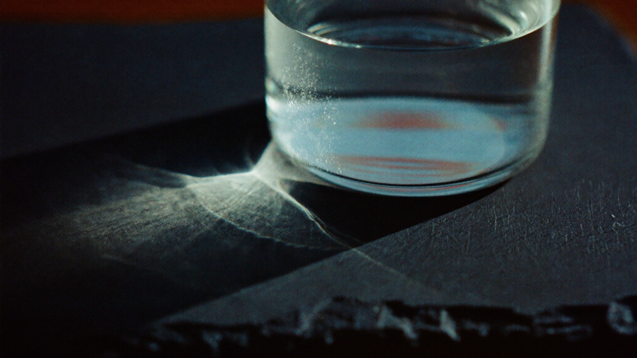 Cool daylight refracting through a glass of water onto dark slate, forming a caustic pattern of blue-teal light